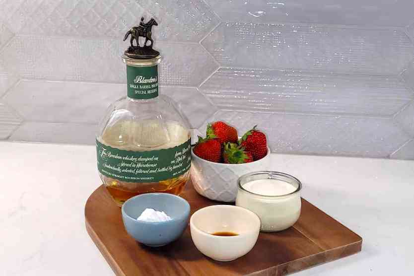 Bourbon, strawberries, whipping cream, sugar and vanilla on a counter.